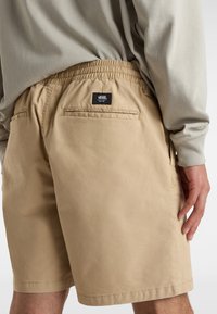 Beige shorts med elastisk talje, med en baglomme og en Vans logo etikette. Materialet ser glat og let ud.