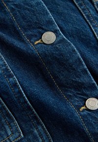Blauer Denim-Stoff mit zwei Metallknöpfen, die "NEXT DENIM SINCE 1982" eingraviert haben, und sichtbaren beigefarbenen Nähten entlang der Nähte.