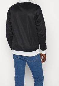 Svart bomberjacka med en slät textur, vita ribbade mudd och fåll, samt en avslappnad passform. Bärs med blå denimjeans.