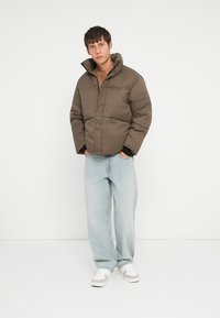 Bruine pufferjack met ruitpatroon en hoge kraag, voorzien van een frontzipper en twee zijzakken, gedragen over een beige trui en lichte jeans.