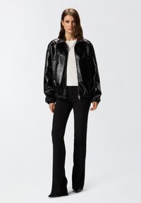 Chaqueta negra brillante con cremallera, con puños elásticos, llevada sobre una camiseta blanca, combinada con pantalones negros ajustados y zapatos de punta negros.