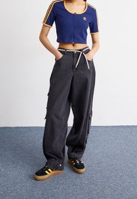 Person som bär en blå kort zip-up-topp med gula ränder, lösa svarta cargo-byxor med ett rep-bälte och svarta sneakers med gula ränder.