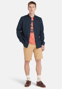 Camicia blu navy, t-shirt grafica arancione, pantaloni corti beige, calzini color crema e scarpe da barca marroni. Outfit estivo casual con colori sovrapposti.