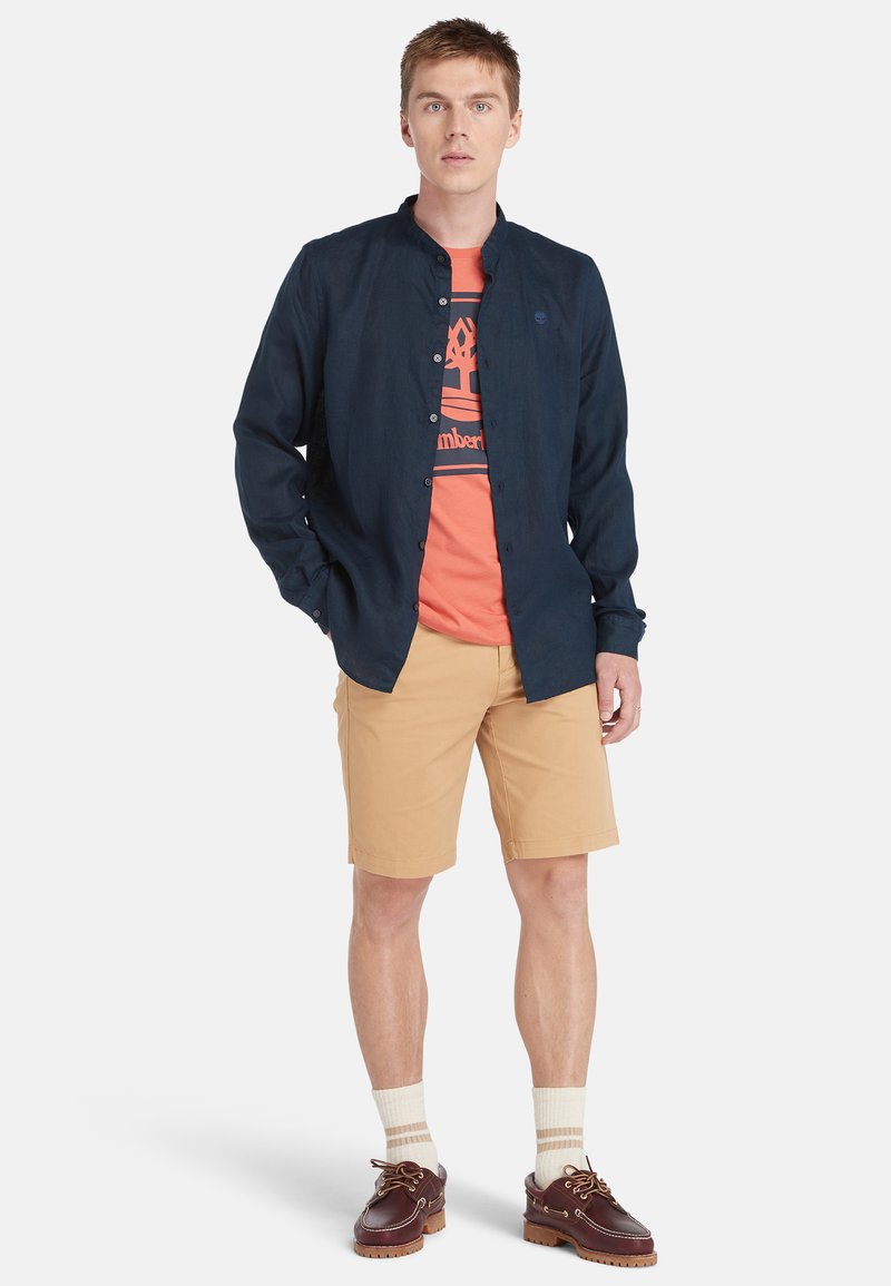 Camicia blu navy, t-shirt grafica arancione, pantaloni corti beige, calzini color crema e scarpe da barca marroni. Outfit estivo casual con colori sovrapposti.