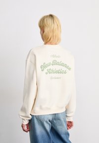 Persoon met kort blond haar, gekleed in een crèmekleurig sweatshirt met de tekst "New Balance Athletics Sportswear" op de achterkant en losse blauwe spijkerbroek.
