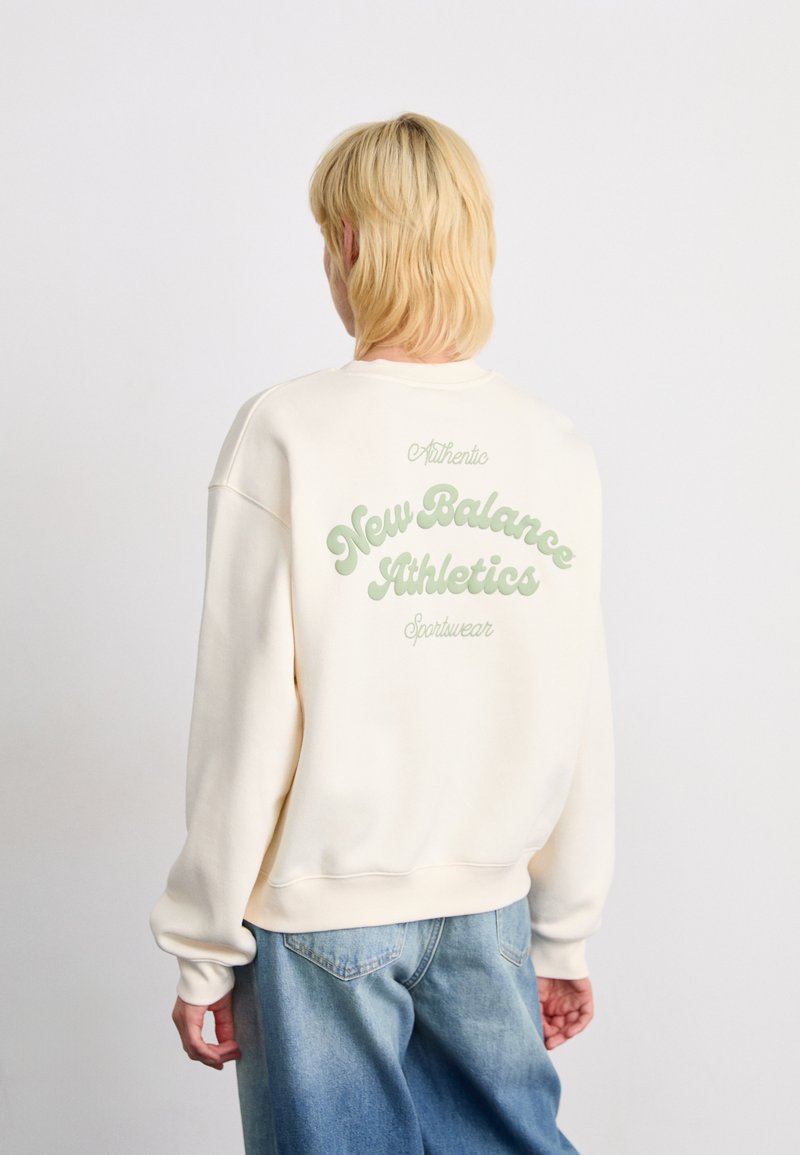 Persoon met kort blond haar, gekleed in een crèmekleurig sweatshirt met de tekst "New Balance Athletics Sportswear" op de achterkant en losse blauwe spijkerbroek.