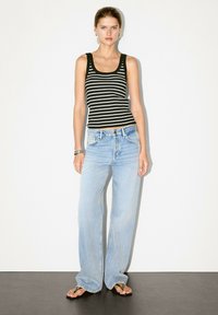 Model staat tegen een witte achtergrond, gekleed in een gestreepte zwart-witte tanktop, lichtblauwe wijde jeans en slippers.