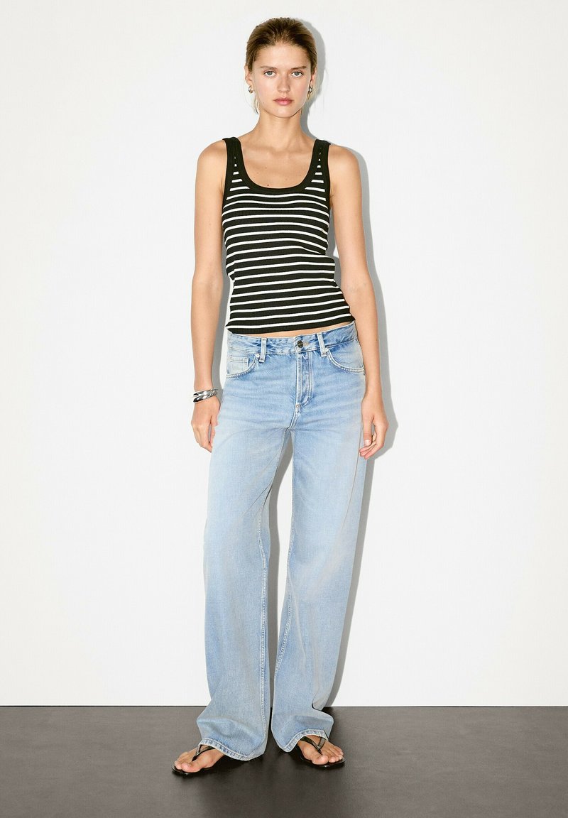 Model staat tegen een witte achtergrond, gekleed in een gestreepte zwart-witte tanktop, lichtblauwe wijde jeans en slippers.