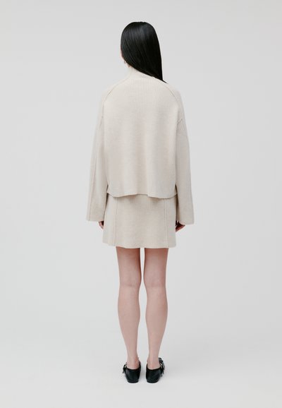 Maglione beige a coste con vestibilità oversize e orlo corto, abbinato a una mini gonna coordinata. Modello che indossa scarpe nere, visto da dietro.