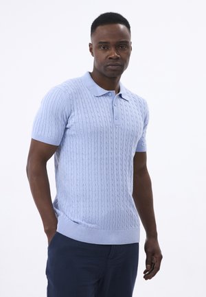 Pull en tricot polo à manches courtes bleu clair avec un motif en chevrons, doté d'un col et de poignets et ourlet ribbés, porté avec un pantalon sombre.