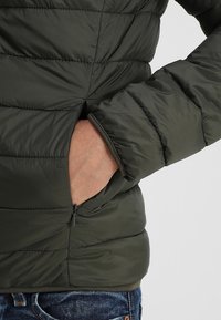 Doudoune vert foncé avec poches latérales zippées, sections matelassées horizontales et un matériau élégant à texture lisse.