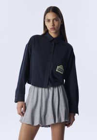 Marinebluse mit Knopfleiste, hochgekrempelten Ärmeln und einer aufgesetzten Tasche mit grüner Stickerei. Kombiniert mit einem hellgrauen, weit ausgestellten Rock.