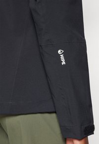 Veste en tissu noir avec une finition texturée, comportant un logo "HIPE" blanc sur la manche et des accents en Velcro au niveau du poignet.