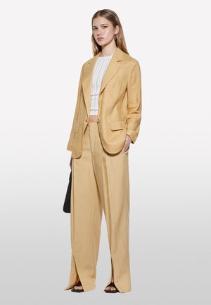 Blazer giallo chiaro su misura e pantaloni a gamba larga in lino. Top corto a coste bianco sotto. Borsa a spalla intrecciata nera. Sandali con la punta aperta.