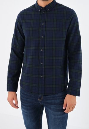 Man draagt een donkerblauw-groen geruit overhemd met knopen en lange mouwen en donkerblauwe spijkerbroek, staand tegen een witte achtergrond.