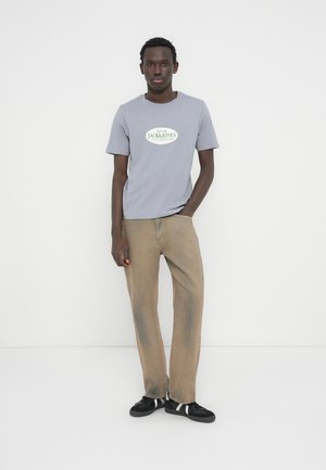 Jauns vīrietis stāv pie baltas sienas, valkājot gaiši pelēku Jack & Jones T-kreklu, izbalējušas bēša krāsas džinsus un melnas sporta kurpes ar baltām svītrām.