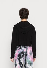 Sweat à capuche noir raccourci avec une texture côtelée et une capuche attachée, associé à un pantalon tie-dye multicolore présentant des motifs roses, noirs et blancs.