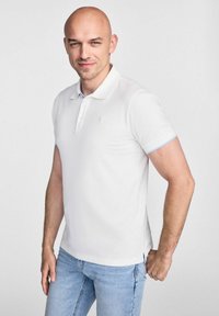 Biała koszulka polo o gładkim wzorze, z jasnoniebieskimi akcentami na rękawach i małym logo po lewej stronie klatki piersiowej, zestawiona z jasnoniebieskimi dżinsami.