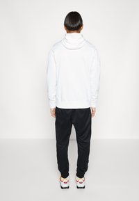 Biała bluza z kapturem i wysokim kołnierzem, zestawiona z czarnymi dresami. Strój posiada elastyczne mankiety i pas, noszony z wielokolorowymi sneakersami.