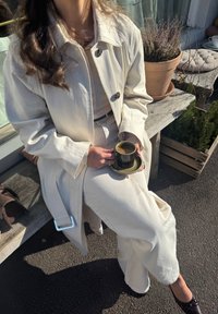 Biała, oversize'owa kurtka z dużymi guzikami i paskiem, w połączeniu z dopasowaną kremową bluzką oraz szerokimi spodniami. Trzyma ciemną filiżankę kawy na talerzyku.