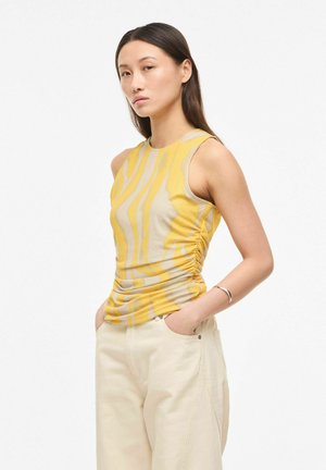 Jeune femme aux longs cheveux noirs portant un haut rayé jaune et beige sans manches et un pantalon beige clair, les mains dans les poches.