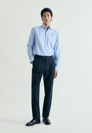 Ung mand iført en lyseblå skjorte med knapper, sorte bukser og sorte loafers, stående mod en ensfarvet lys baggrund.