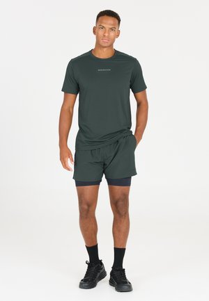 Donkergroen sportshirt met korte mouwen, gecombineerd met bijpassende shorts. Heeft een minimaal logo op het shirt. Zwarte sneakers en sokken.