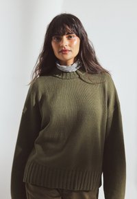 Khaki green knitted sweater with ribbed hem and crew neckline. Model wears a light gray turtleneck underneath, showcasing layered texture.