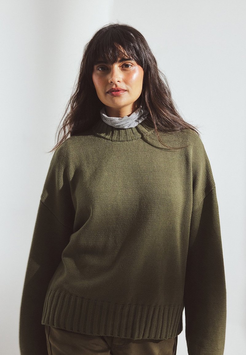 Khaki green knitted sweater with ribbed hem and crew neckline. Model wears a light gray turtleneck underneath, showcasing layered texture.