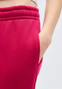 Κοντινό πλάνο ενός ατόμου που φοράει φωτεινά ροζ παντελόνια φόρμας με ελαστική μέση και το χέρι χαλαρά τοποθετημένο στην πλαϊνή τσέπη.