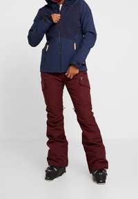 Giacca da neve blu navy con zip frontale, tasche e polsini elasticizzati; abbinata a pantaloni da neve bordeaux con zip e tasche laterali.