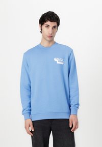 Gaiši zila kokvilnas maisījuma būtne, ar apaļu kaklu, gar�ām piedurknēm un baltu "TOMMY JEANS" logotipu uz krūtīm.