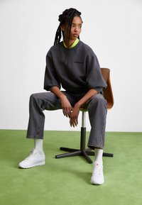 Grauer übergroßer Sweatshirt mit Logo, grüner Kragen, graue Jeans und weiße Sneakers. Geflochtenes Haar, sitzt auf einem Holzstuhl vor einem grünen Teppich.