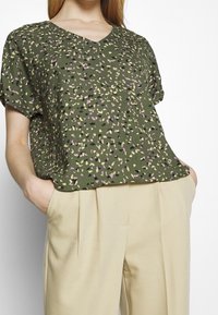 Top verde a maniche corte con scollatura a V, caratterizzato da un motivo maculato in viola, giallo e nero. Abbinato a pantaloni beige plissettati.