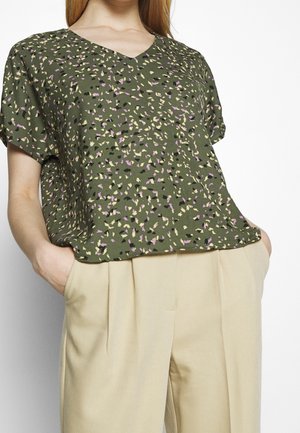Top verde a maniche corte con scollatura a V, caratterizzato da un motivo maculato in viola, giallo e nero. Abbinato a pantaloni beige plissettati.