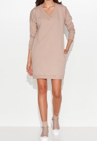 Beżowa sukienka dresowa z długim rękawem, wycięciem w kształcie litery V, bocznymi kieszeniami i prążkowanym dołem. Połączona z jasnoszarymi tenisówkami za kostkę.