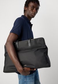 Fiatal férfi tengerészkék pólóban és farmerban, egy nagy fekete bőrtáskával a rövid fogantyúkkal, egy sima háttér előtt.