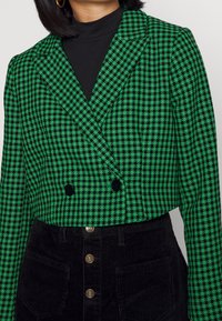 Zeleno-černé sako se vzorem psí zuby a dvouřadým zapínáním, zdobené černými knoflíky, doplněné černou manšestrovou sukní a vysokým límcem.