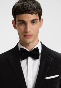 Zwarte fluwelen tuxedo-jack met satijnen revers, gecombineerd met een wit overhemd en een zwarte fluwelen vlinderdas; voorzien van een witte pochet.