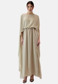 Beige, vloeiende jurk met een lijfje dat bij de taille samenkomt, draperende cape-mouwen en een gladde, lichte stof. Zwarte sandalen gedragen.