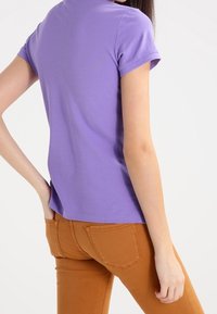 Fialová košile s krátkým rukávem a klasickým límcem, vyrobená z hladkého bavlněného materiálu, v kombinaci s úzkými tan kalhotami s kapsami na zadní straně.