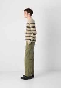Beige striped knit sweater with black lines, paired with olive green pants and black shoes; model stands in profile against a white background.