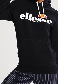 Sweat à capuche noir avec le logo "ellesse" blanc, doté d'une poche kangourou à l'avant, d'une capuche avec cordon de serrage, et de poignets et ourlet côtelés. Associé à un pantalon rayé.