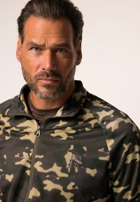 Camouflage zip-up jack in groen, beige en zwart. Beschikt over een hoge kraag en een subtiel logo op de borst, gemaakt van een gladde, textuurstof.