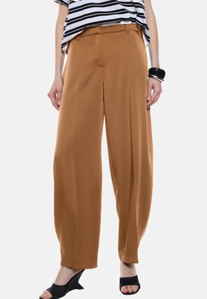 Pantaloni a gamba larga in un caldo tessuto marrone con una texture liscia, caratterizzati da pieghe sul davanti, abbinati a una maglietta a righe e scarpe nere.