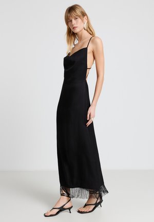 Femme debout portant une robe noire sans manches avec un dos décolleté et un ourlet à franges, des sandales noires à brides et de grandes boucles d'oreilles dorées.