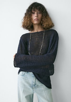 Persona con capelli ricci che indossa un maglione blu navy a maglia larga, jeans azzurri e una lunga collana d'argento, in piedi contro un muro bianco.