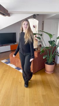 Juodas megztinis ir kelnės vaikščiojantys ant medinių grindų. Šviesiai rudos spalvos tekstūruota sofa su vazoniniu augalu šalia. Šiuolaikinio interjero dizaino elementai.