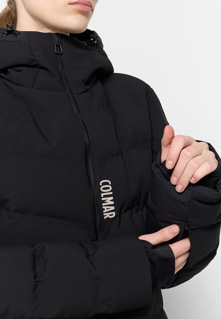 Primo piano di una persona che regola la cerniera del polsino su un giubbotto invernale nero, con il logo "COLMAR" visibile sul davanti.