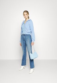 Jovem mulher a usar um hoodie azul claro, uma blusa canelada com botões, jeans azuis, botas brancas até ao tornozelo e a segurar uma pequena carteira azul clara.